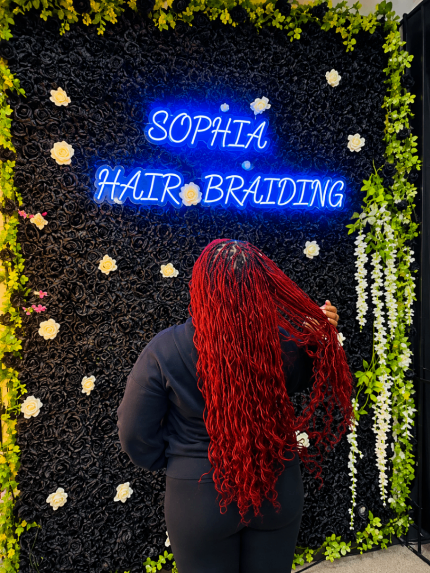 Back view of long red goddess braids with wavy curly ends posed at the Sophia Hair Braiding neon signage wall