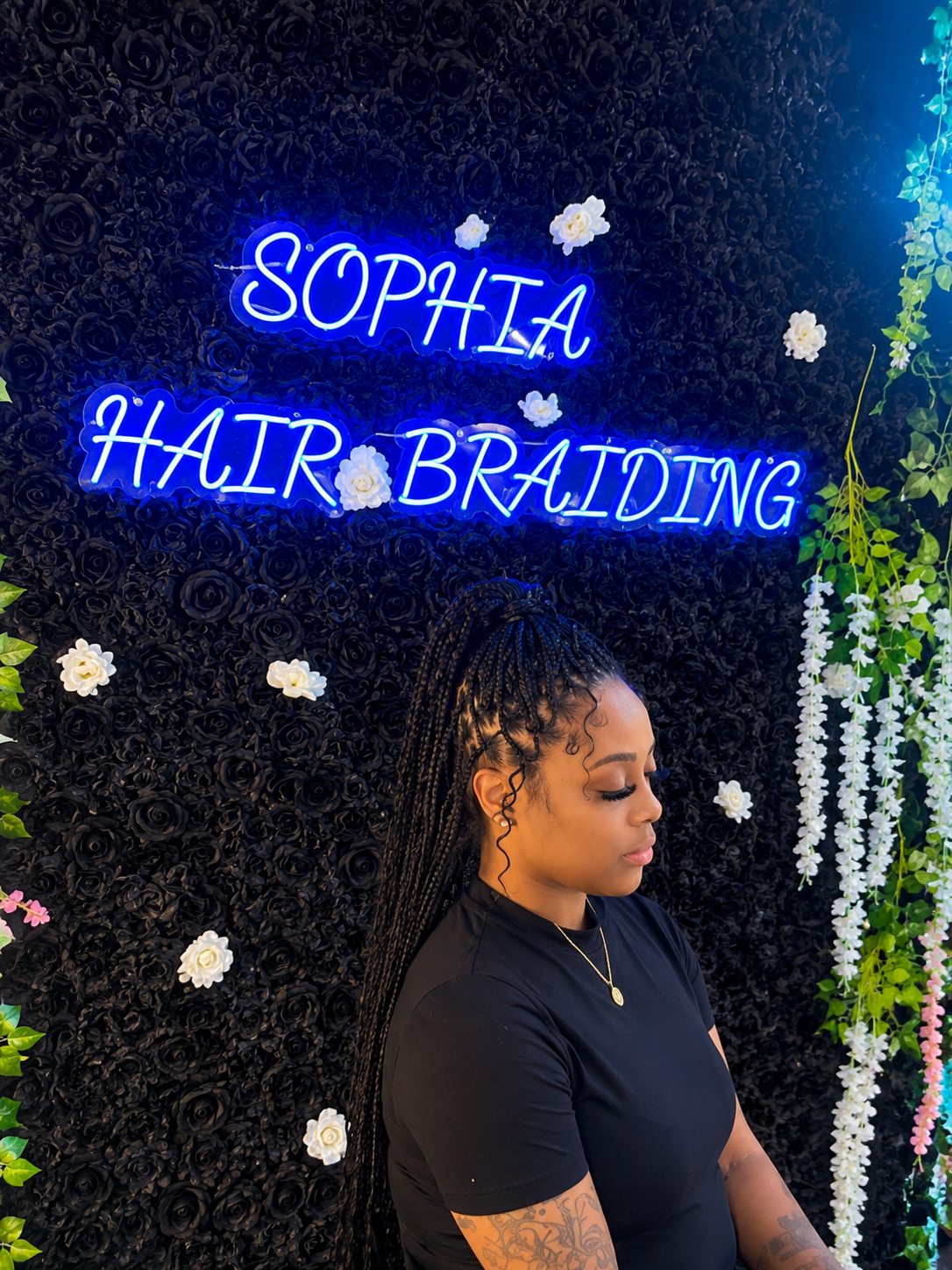 Client with half-up knotless braids and curly baby-hair tendrils posed beneath the blue neon Sophia Hair Braiding sign on the black rose wall