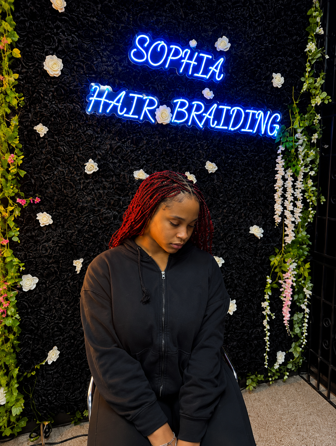 Client with long red twist braids seated in front of the black rose wall beneath the blue Sophia Hair Braiding neon sign