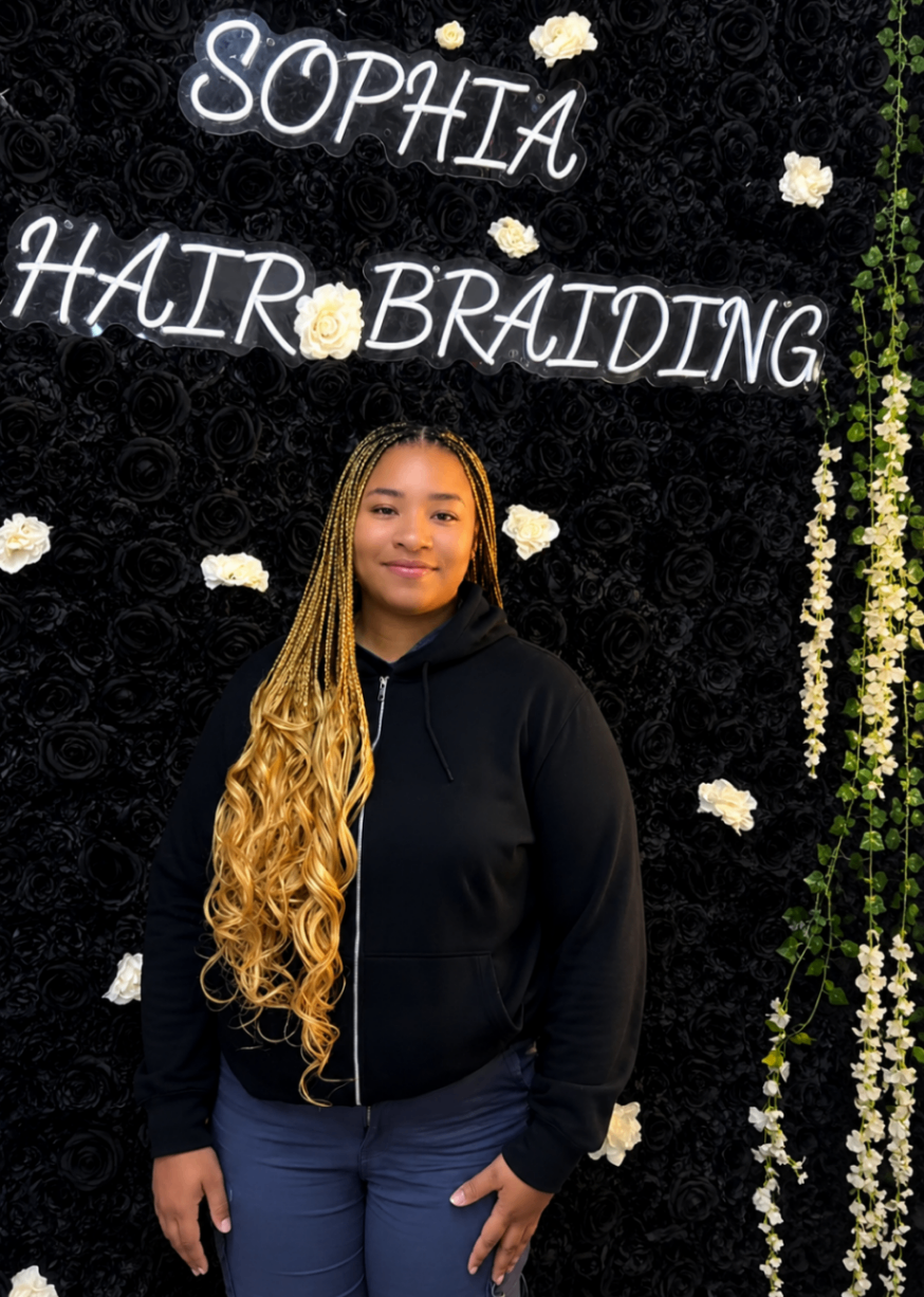 Full-length blonde ombre knotless braids with curly ends posed under the Sophia Hair Braiding neon sign